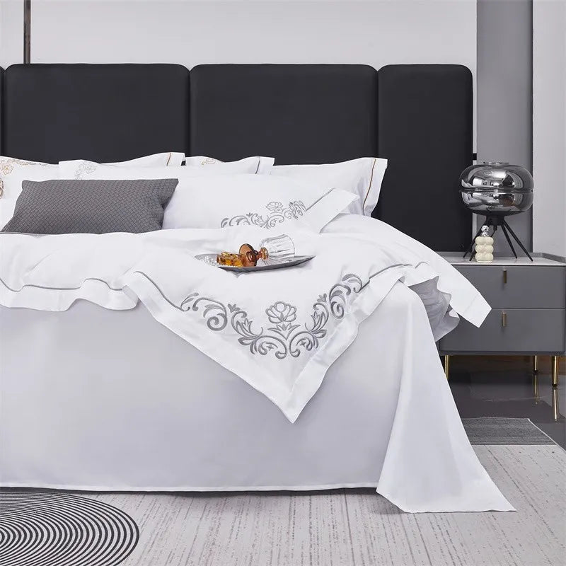 Bed with white bedding featuring intricate embroidery, gray pillows, and a nightstand with decorative items.