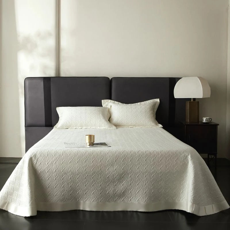 Bedroom with a bed covered in a white quilt, dark headboard, and nightstand with lamp.