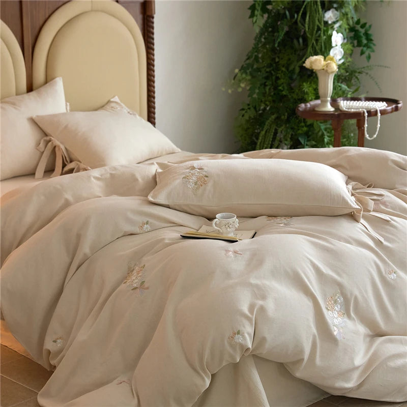 Beige embroidered bedding set on a bed with a side table and plants in the background.