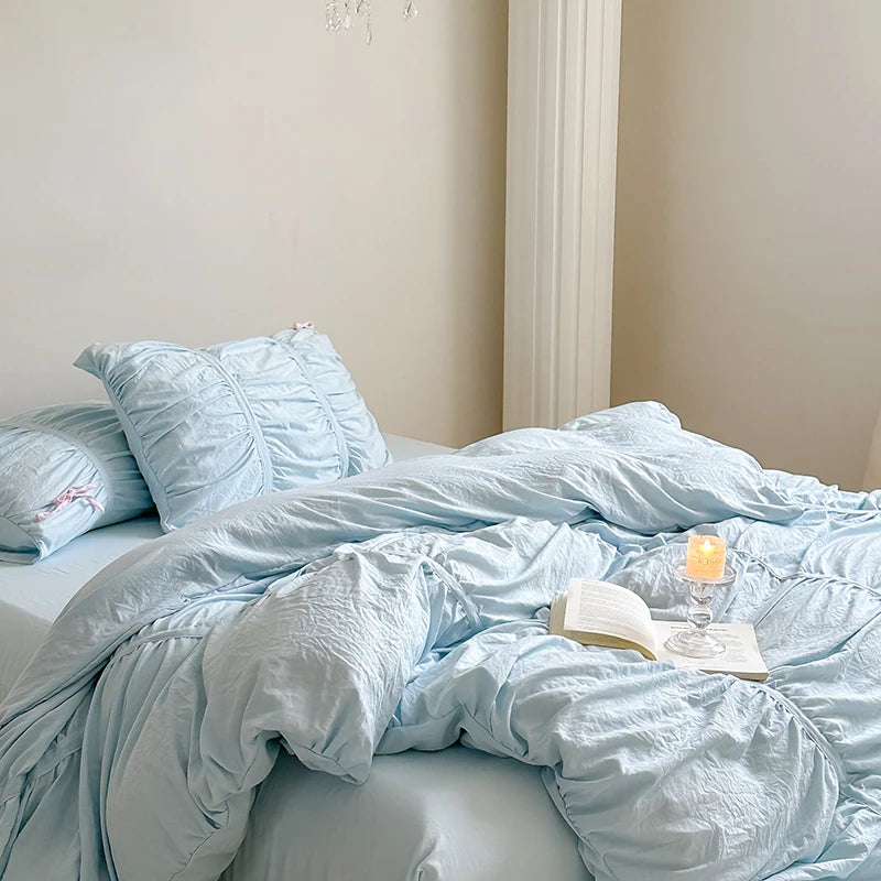 Light blue bedding on a bed with an open book and candle in a glass holder.