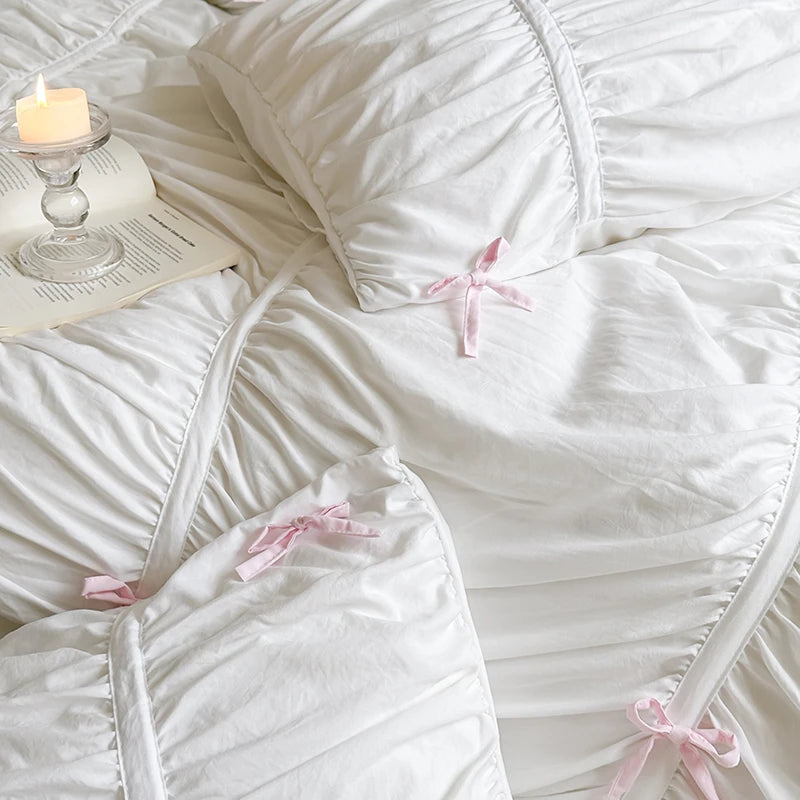 White comforter with pink bows on a bed next to a lit candle and book.