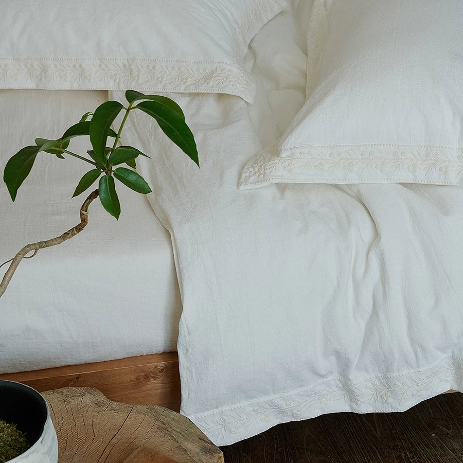 White linen bedding with lace trim on a wooden bed frame, accompanied by a small plant.
